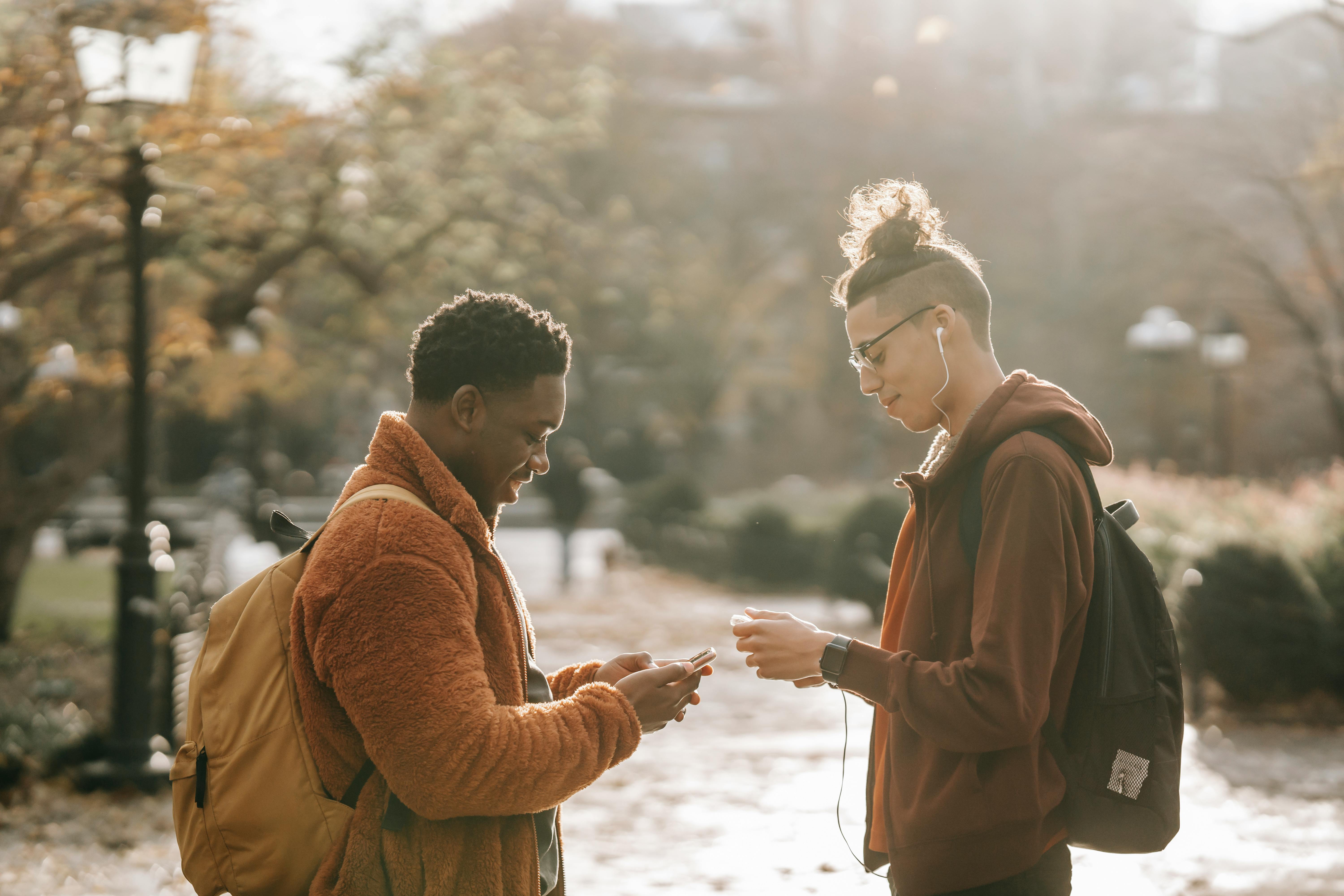 cheerful multiracial friends chatting on smartphones while standing in autumnal park