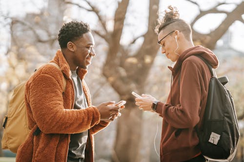 Free Side view of smiling young multiracial male friends wearing warm clothing standing opposite each other and  browsing cellphones on blurred background Stock Photo