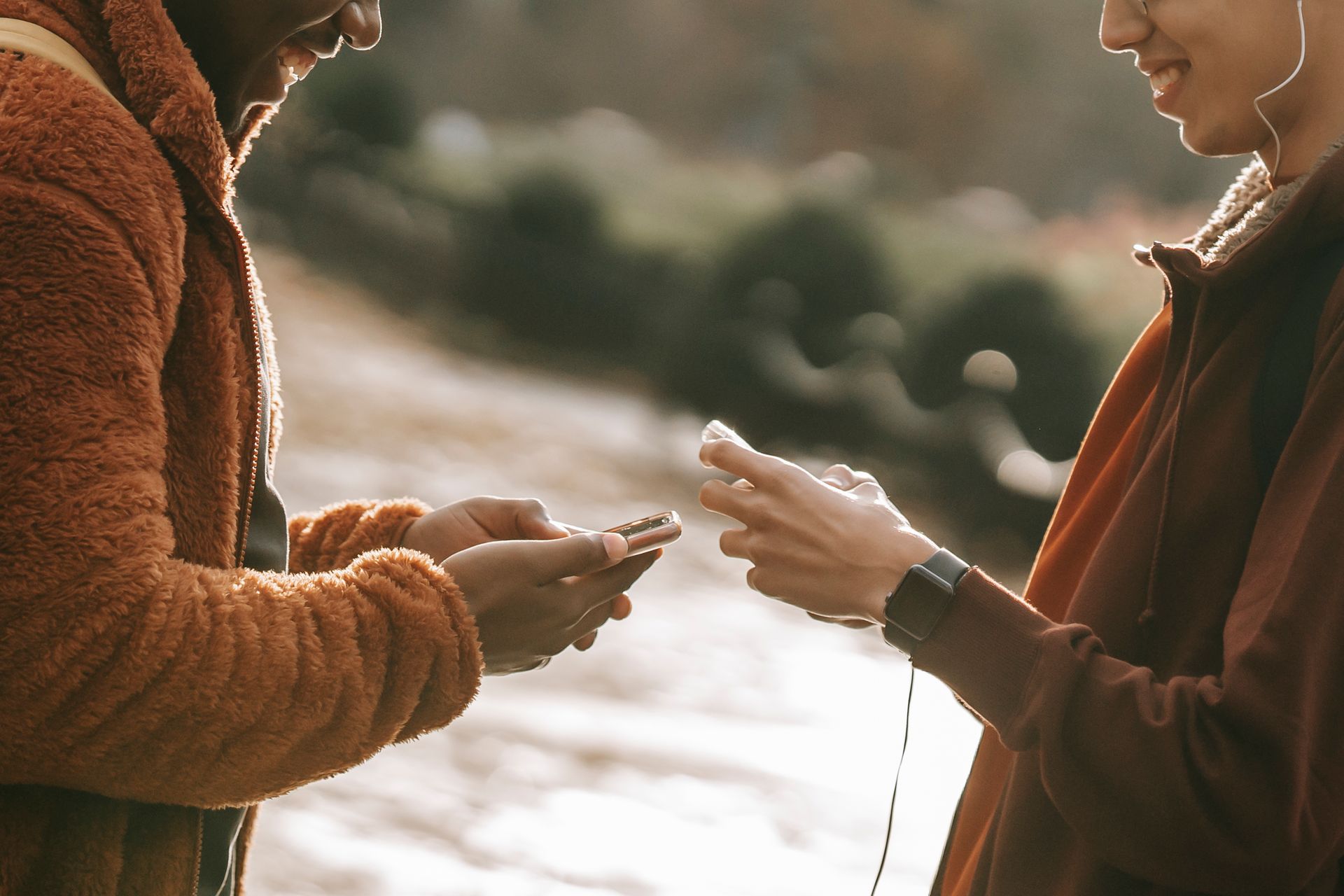 Side view of crop multiethnic guy wearing warm clothes chatting on modern smartphones while standing on blurred background together