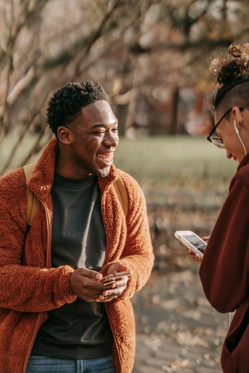 Free Delighted young multiracial male friends wearing warm clothes standing in autumn park and laughing while browsing modern smartphones Stock Photo