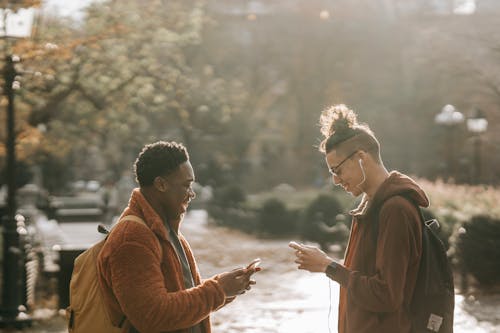 Free Side view of millennial laughing multiethnic friends wearing similar clothes using modern cellphones while standing in city park on autumn day Stock Photo