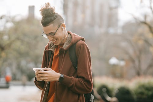 Young ethnic guy with backpack smiling while listening to music on smartphone