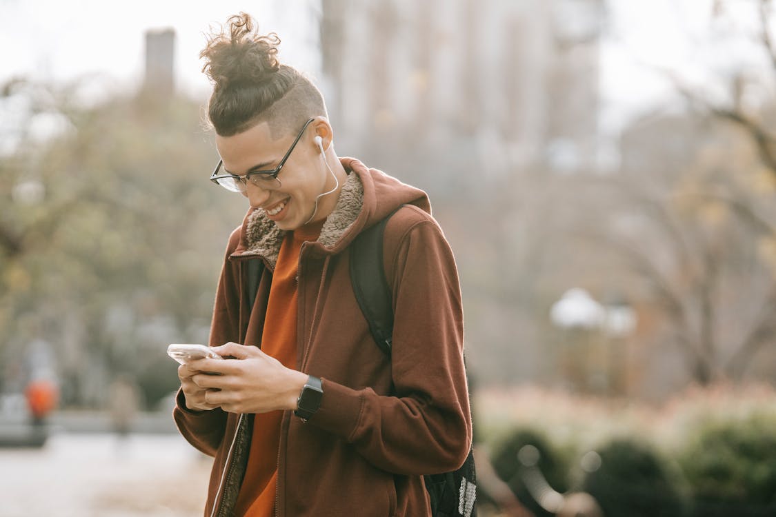 Young ethnic guy with backpack smiling while listening to music on smartphone