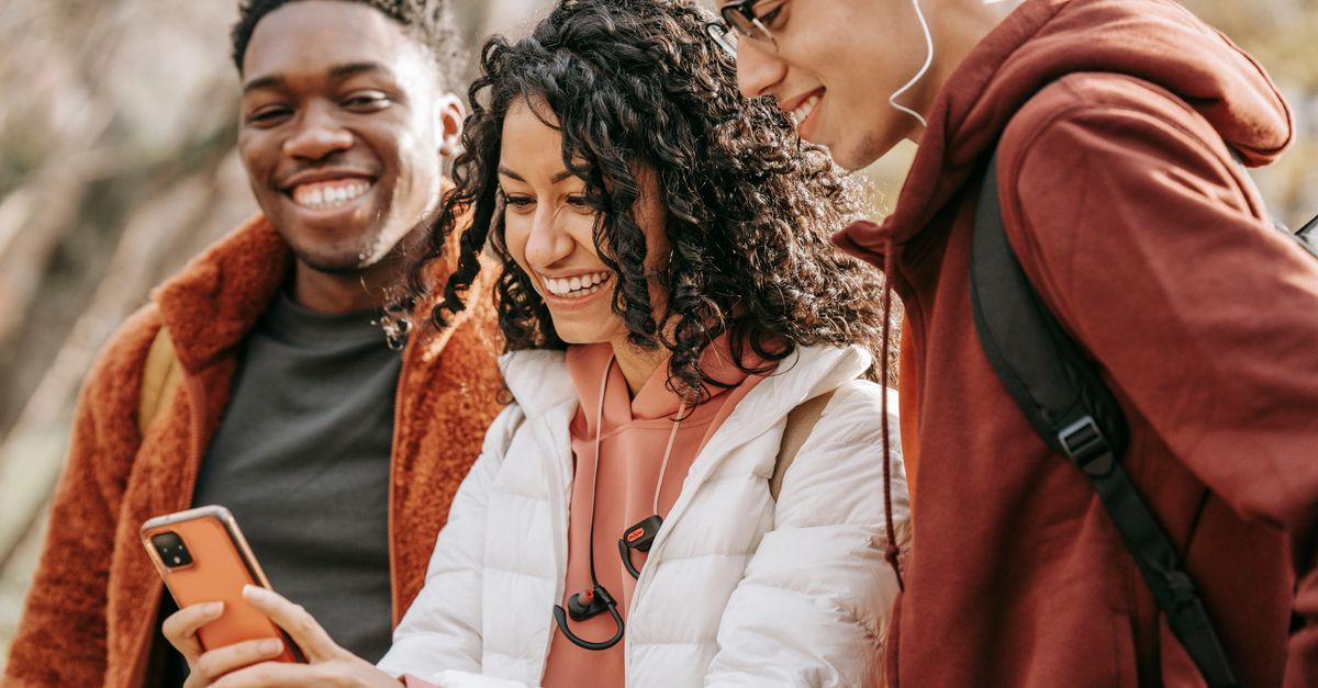 Happy multiracial young friends in casual outfits standing in garden and taking selfie on smartphone while toothy smiling