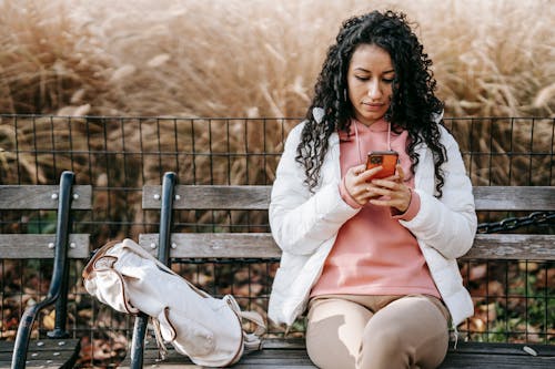 Free Young Latin American female in stylish outfit sitting on bench in fall park while browsing mobile phone and looking at screen Stock Photo