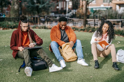 Free Full body of multiracial classmates in trendy clothes sitting on grass and studying with notepads and laptop Stock Photo