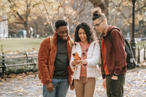 Free Group of diverse friends in stylish outfits standing in fall park with smartphone and taking selfie while smiling and looking at screen Stock Photo