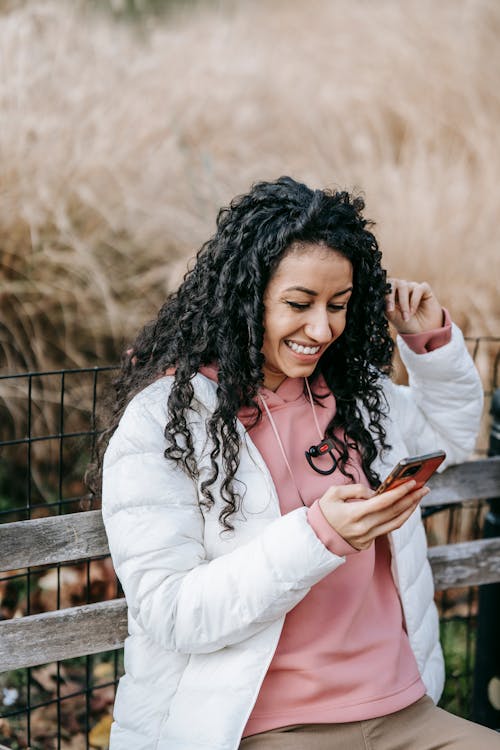 Cheerful Latin American female with curly hair browsing mobile phone while sitting on wooden bench and looking at screen