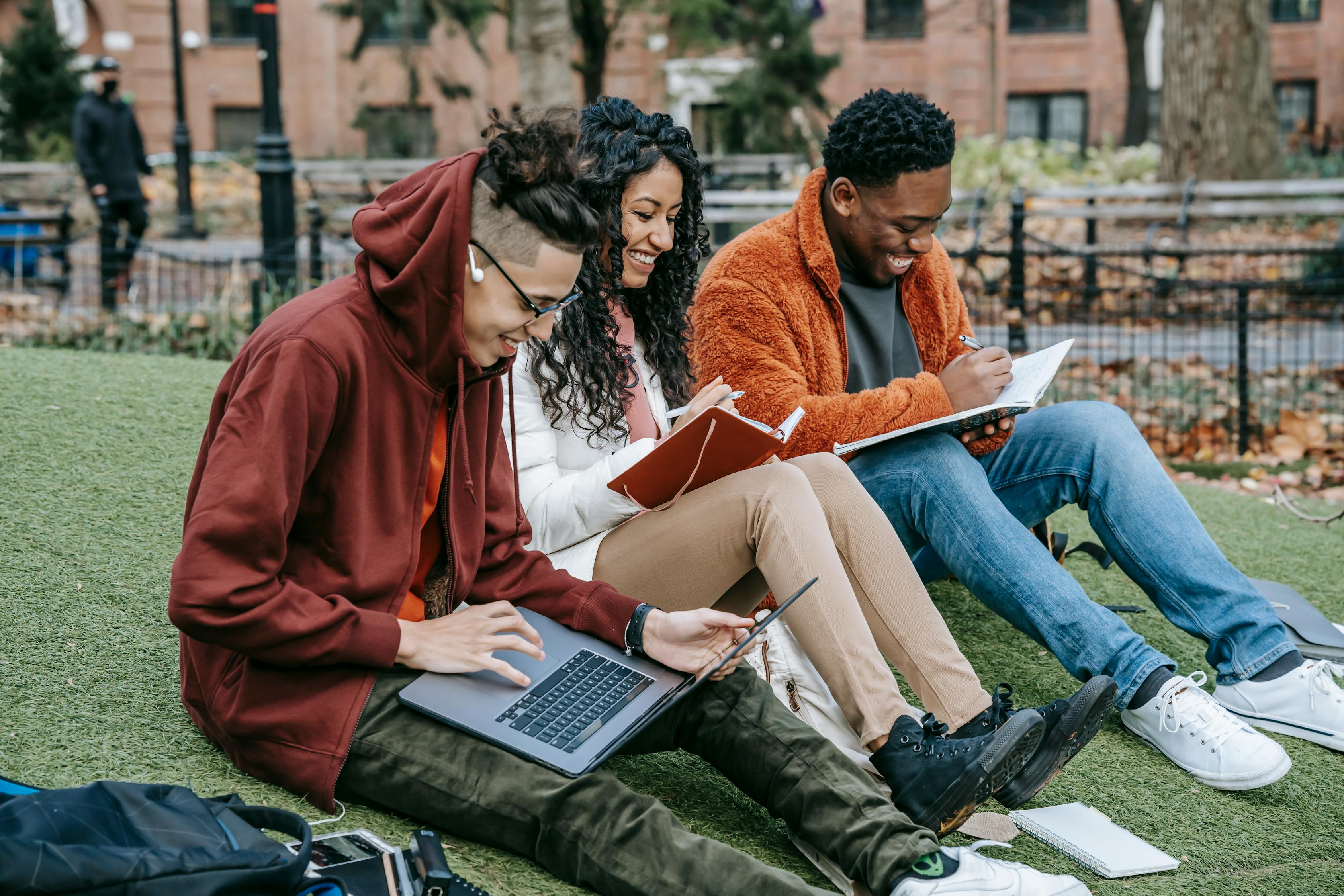 Happy multiracial group of students in casual clothes sitting together on grass and studying with laptop and books