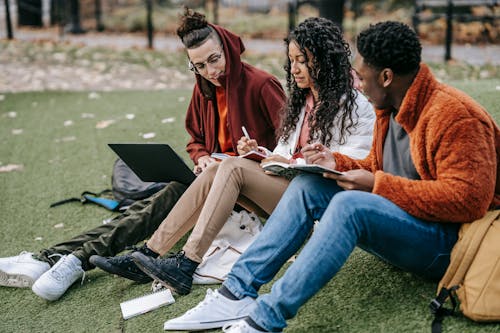 Young multiracial students in trendy clothes sitting on grass and studying together in autumn park