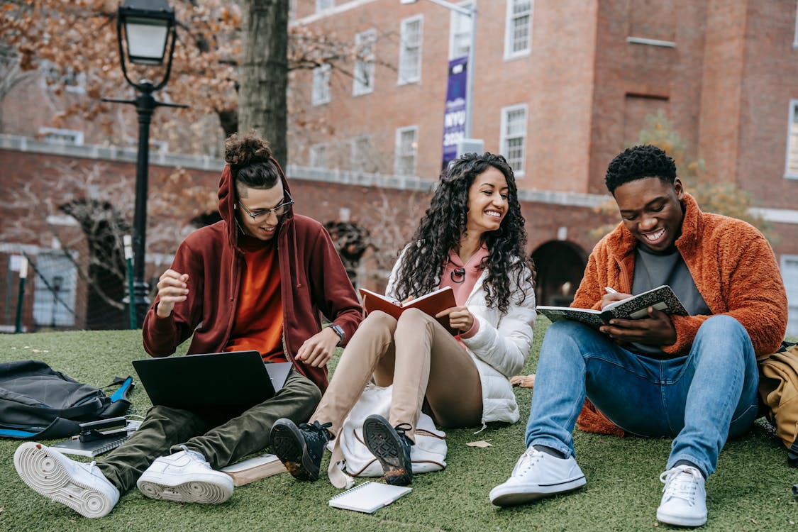 Free Full body of happy diverse students with notebooks and laptop sitting on grassy lawn on campus of university while studying together Stock Photo