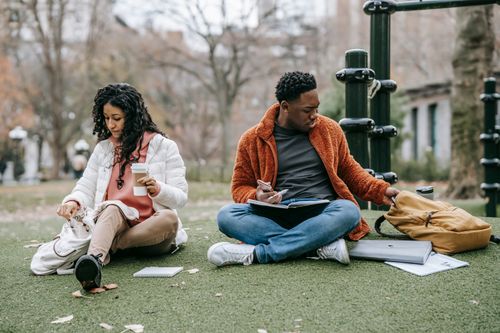 Free Full body of ethnic woman with takeaway coffee and black man with notebook opening rucksacks while sitting on grassy ground during studies together Stock Photo