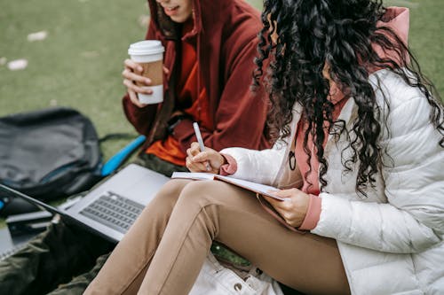 Side view of unrecognizable classmates with takeaway coffee browsing laptop and taking notes in notebook while preparing for classes on grassy ground