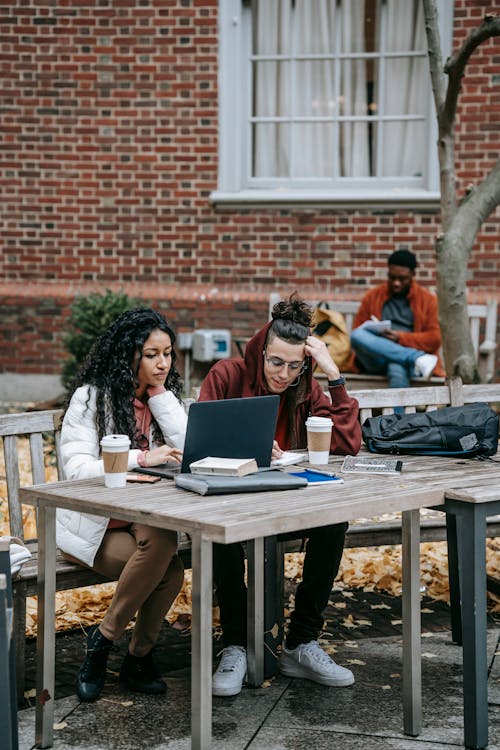 Full body of multiethnic students surfing netbook at table with takeaway coffee while studying together on street with distant black friend