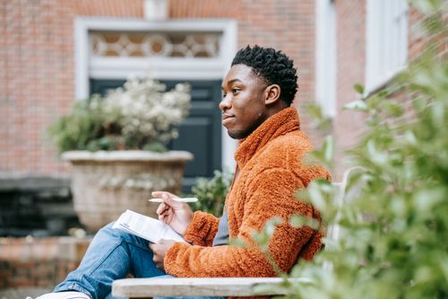 Free Side view of thoughtful African American male student writing on paper while sitting on bench near building on street during homework preparation on blurred background Stock Photo