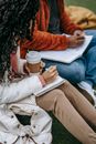 Unrecognizable diverse diligent classmates with takeaway coffee taking notes in copybook while sitting on lawn during studies preparation on street