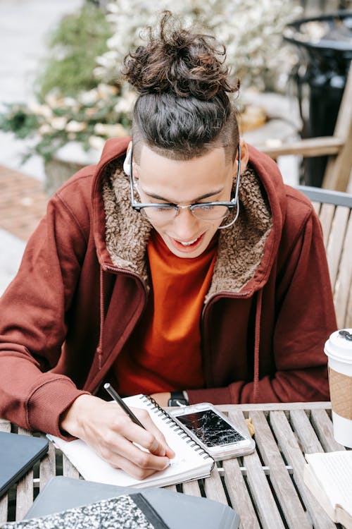 Focused male student with eyeglasses taking notes in notepad while sitting at wooden table with smartphone on street while doing homework