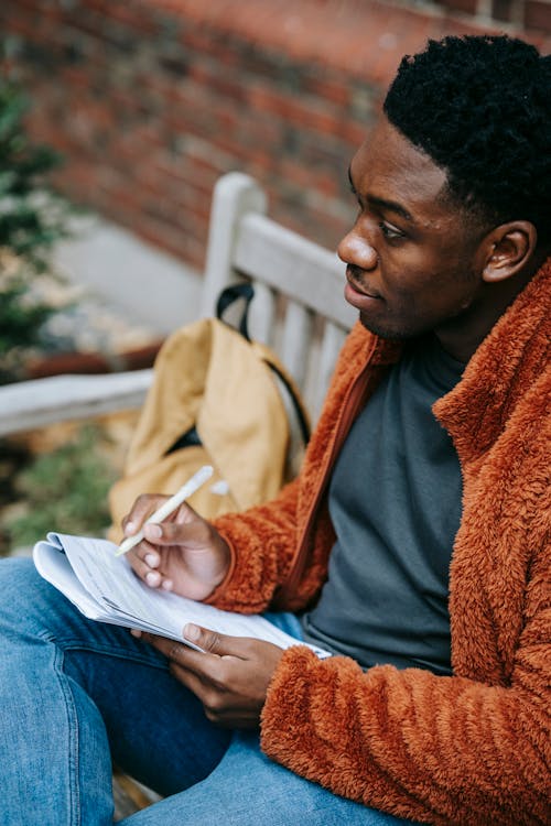 From above of crop African American male in warm clothes writing in organizer while looking away on blurred background