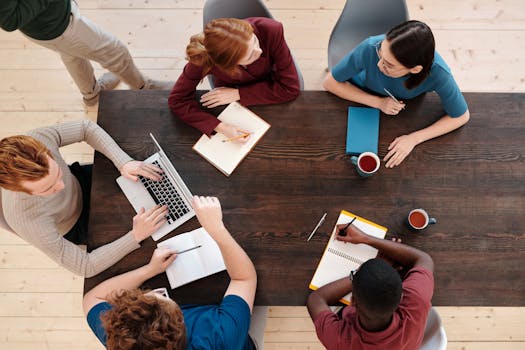 A group of diverse colleagues collaborate in a modern office setting around a wooden table.