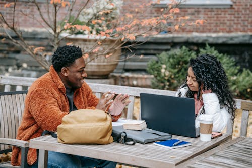 Cheerful multiracial students speaking and smiling at table with coffee and laptop on street