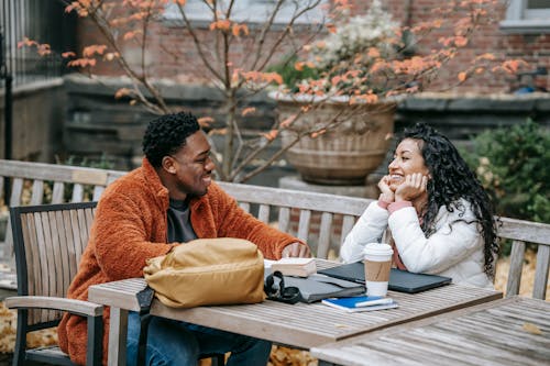 Happy diverse young friends in casual outfits speaking at table with laptop and coffee in yard