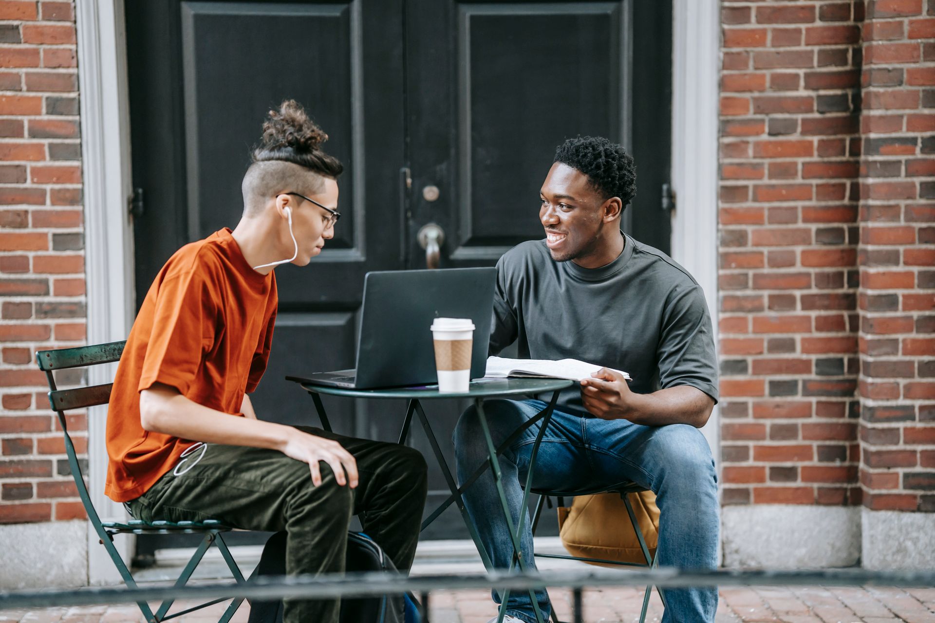 Cheerful multiracial friends discussing university at table with netbook and coffee near entrance of brick building