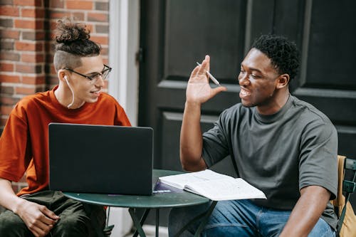 Cheerful young multiethnic male friends smiling and chatting while sitting together in street cafe and doing homework assignment using laptop