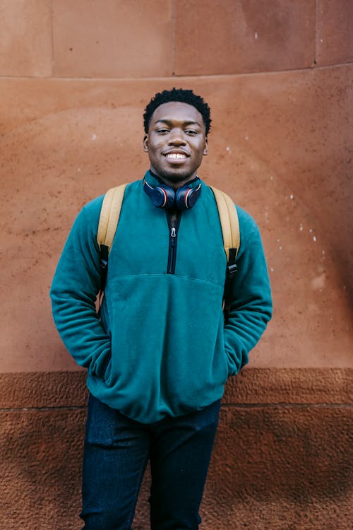 Positive young African American male student in casual clothes and backpack standing near stone building with hands in pockets and smiling