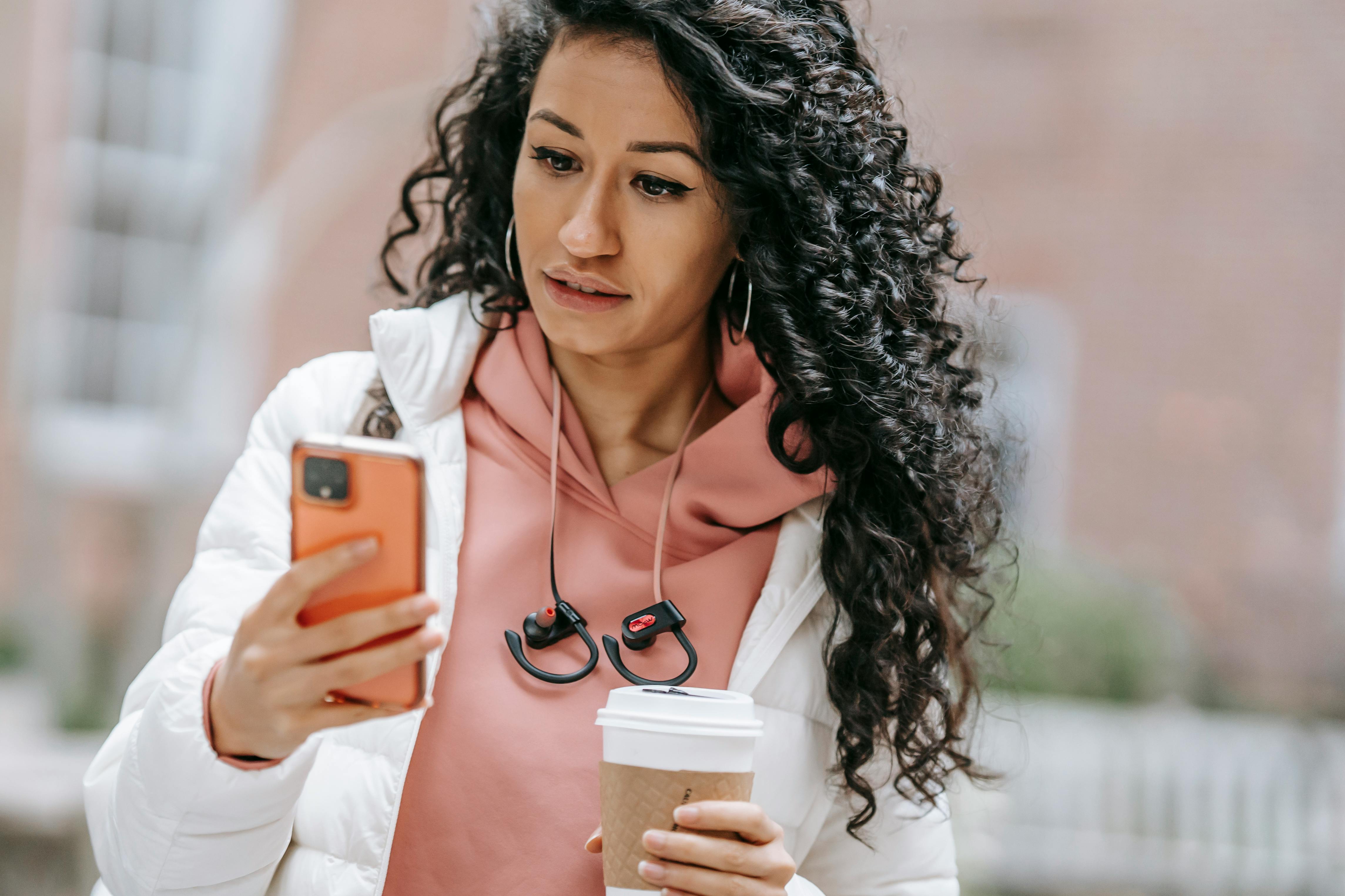 serious young ethnic woman using smartphone and drinking coffee to go on street