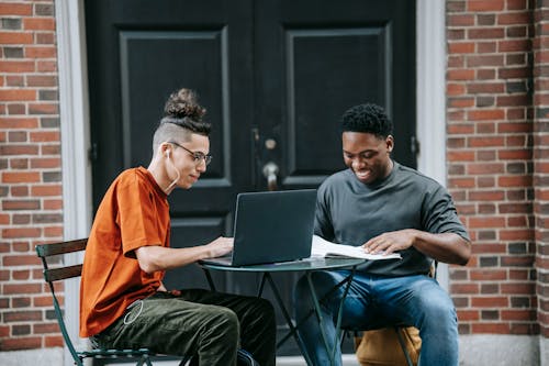 Positive young diverse male colleagues in casual clothes smiling and using laptop while working remotely on project sitting at table in street cafe