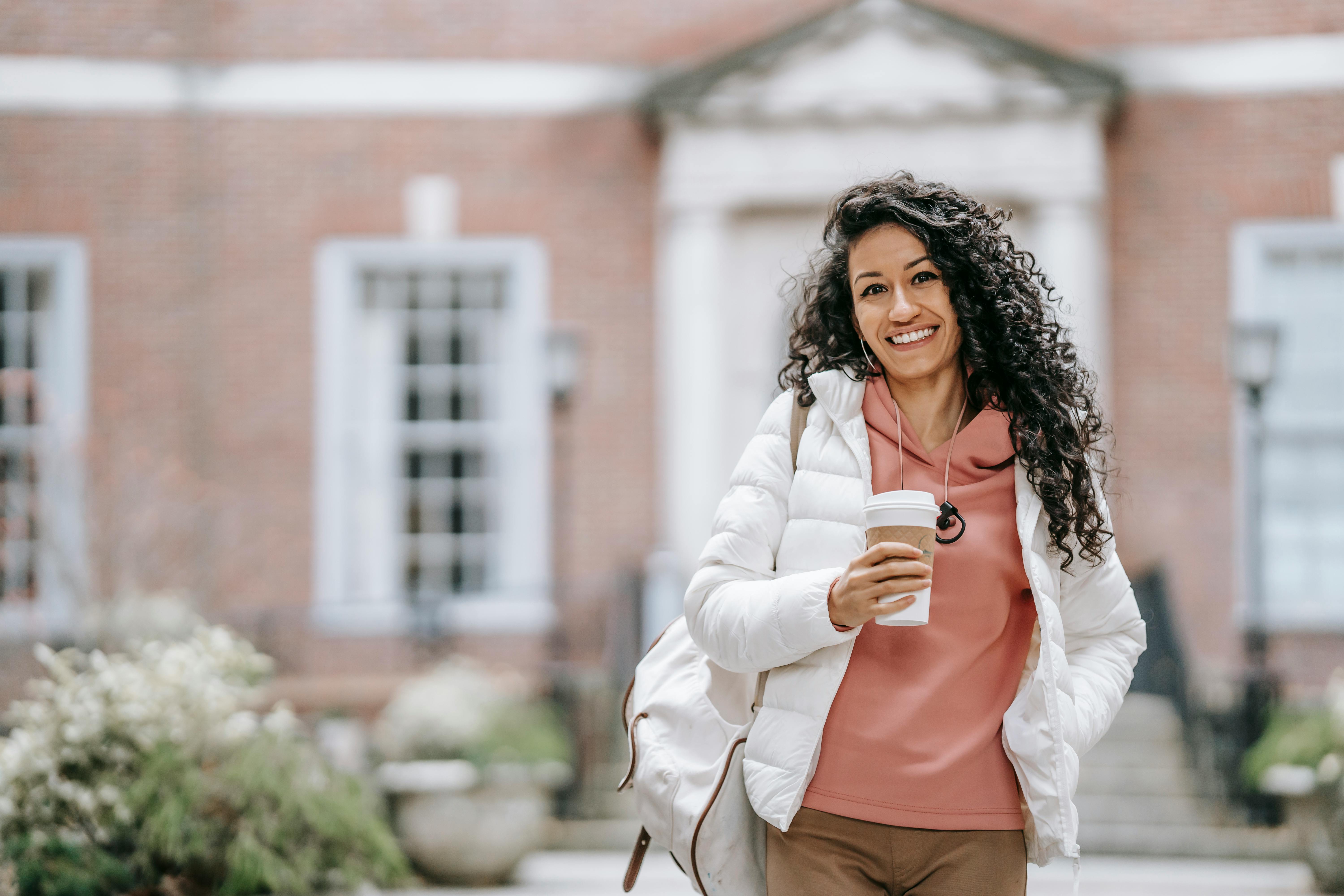 cheerful young ethnic woman drinking takeaway coffee and smiling near brick building