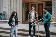 Young ethnic outcast female walking on street near diverse bullying male classmates standing on building stairs and smiling