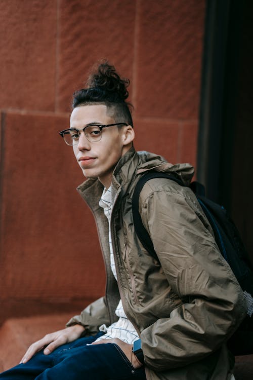 Side view of young calm ethnic male student in stylish outfit and eyeglasses sitting on stone stairs on city street and looking away