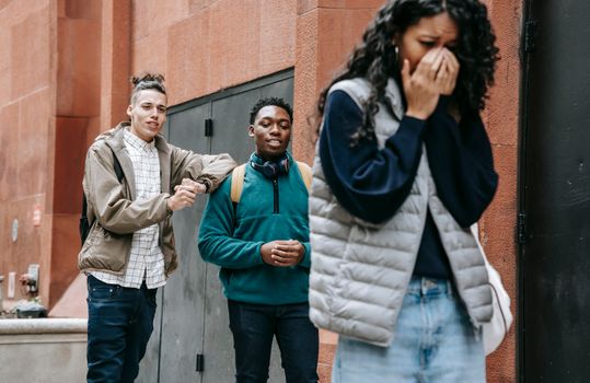 Low angle of annoyed young ethnic lady covering mouth and crying after being abused by rebellious multiracial male groupmates