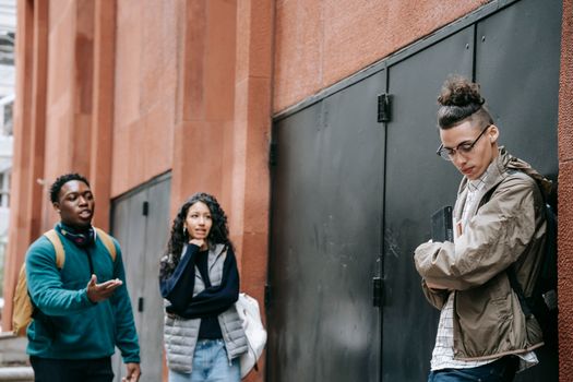 Low angle of aggressive young diverse groupmates bullying shy upset male leaning on building and looking down