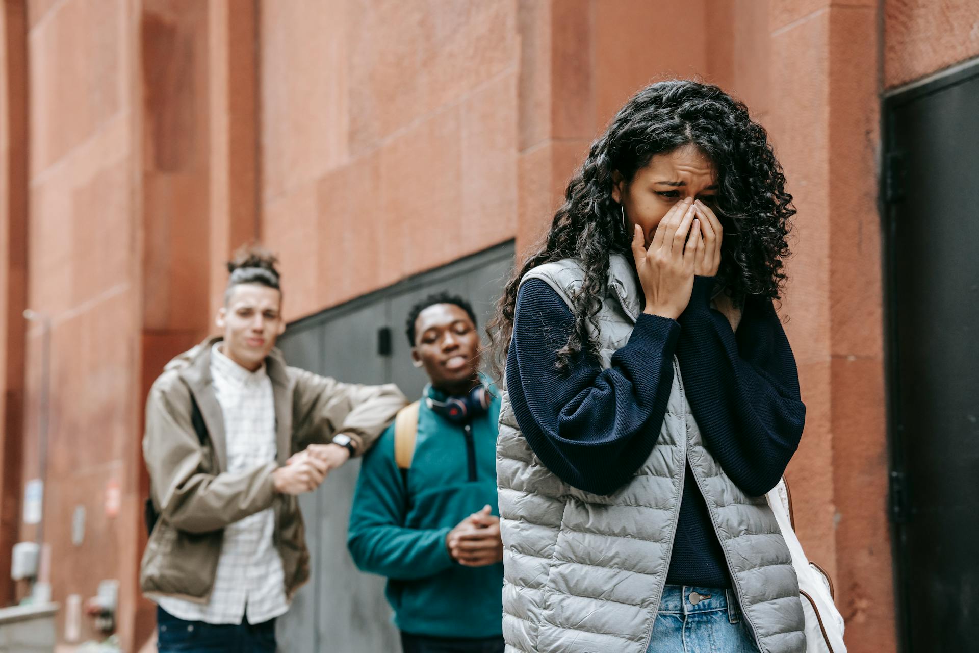 From below of despaired young ethnic female student covering mouth with hands while crying on street after being bullied by multiracial classmates