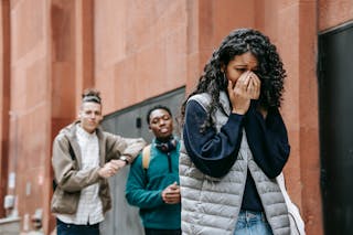 From below of despaired young ethnic female student covering mouth with hands while crying on street after being bullied by multiracial classmates