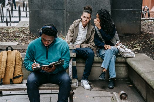 Young multiethnic friends gossiping about black male student preparing for exams in park