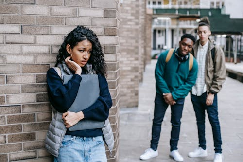 Free Latin American woman leaning on brick wall while hiding from multiracial classmates in college Stock Photo