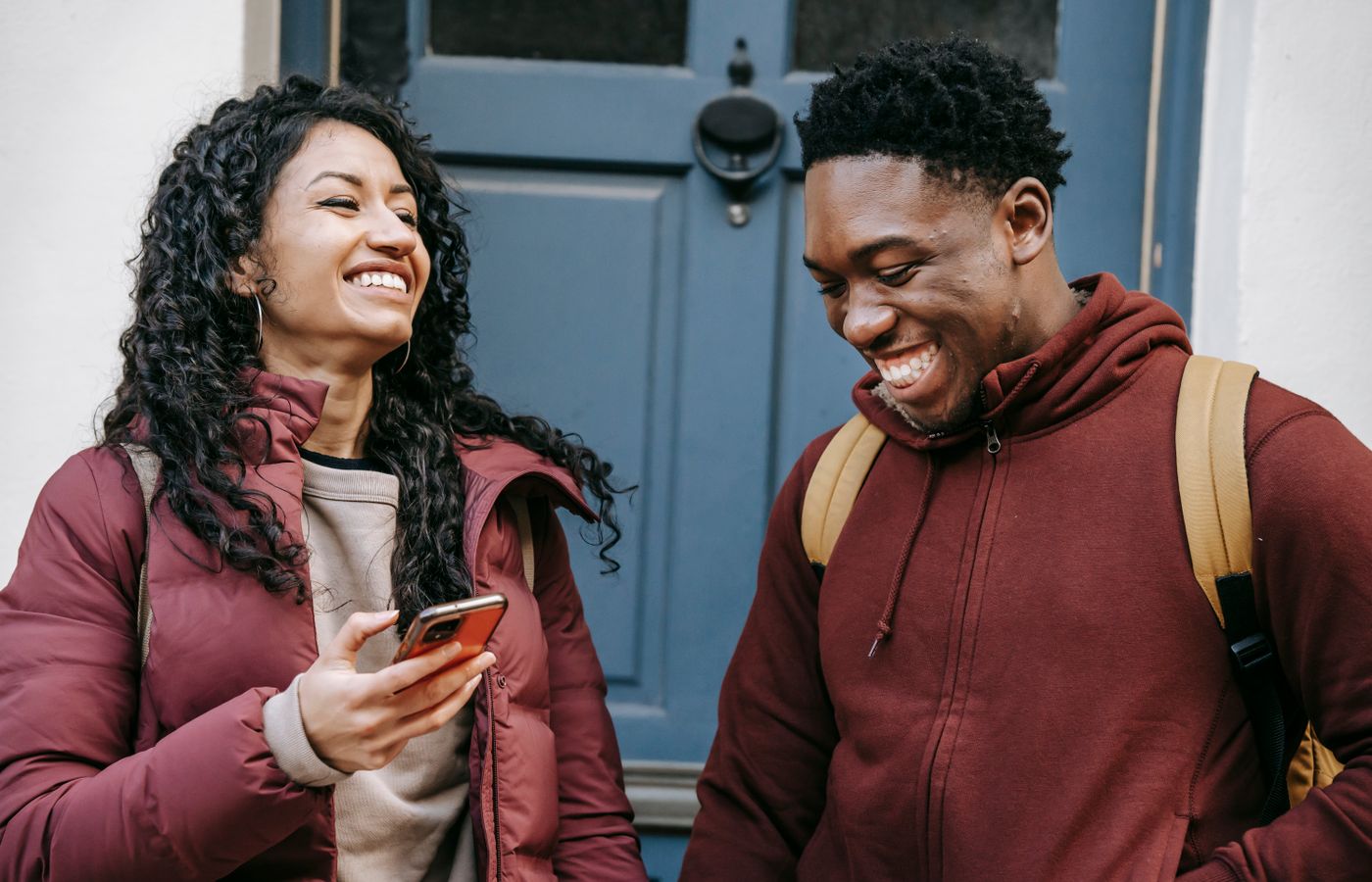 Two young adults smile and chat in front of a blue door, enjoying a light moment outdoors.
