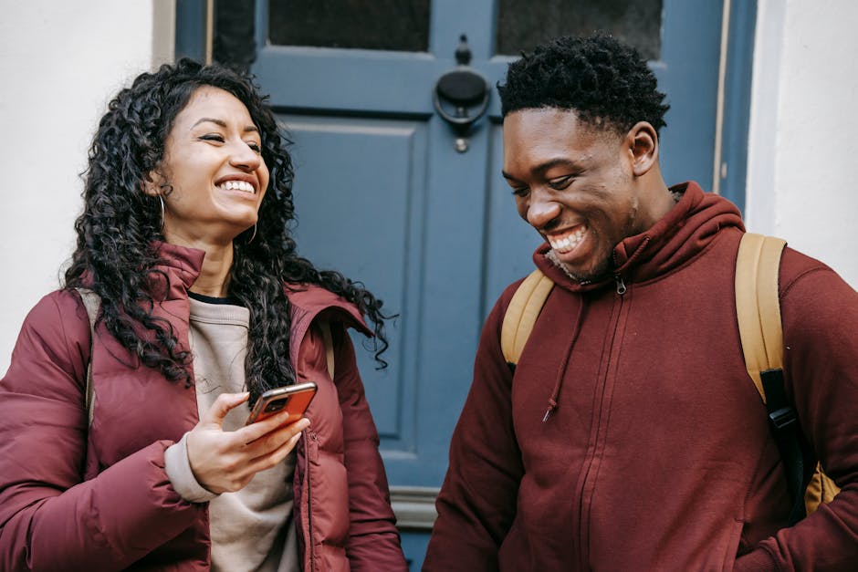 Diverse cheerful friends with smartphone laughing near door of building