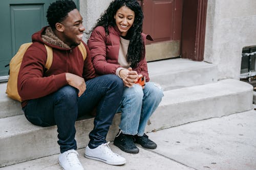 Free Cheerful multiethnic crop couple laughing and smiling while holding hands and resting on concrete steps Stock Photo