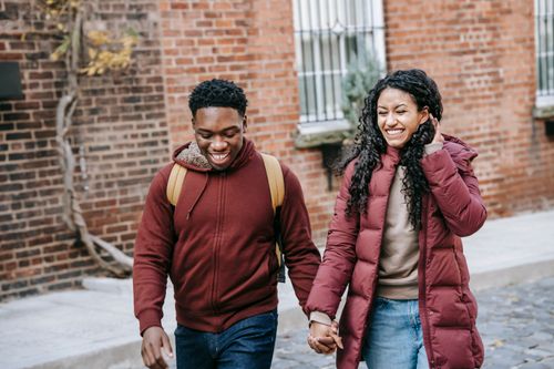 Free Multiethnic young glad couple holding hands and smiling while promenading near brick building on urban street Stock Photo