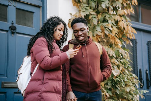 Delighted young multiracial couple using smartphone and holding hands near door of building with verdant leaves