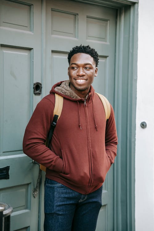 Cheerful young African American male with hands in pocket smiling and looking away near door of house