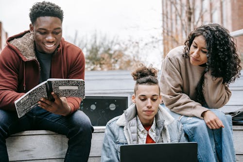 Group of multiethnic happy young students doing homework with copybook and netbook on street