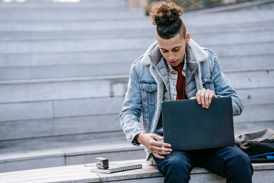 Concentrated male with stylish hairstyle browsing netbook while sitting on wooden stairs with notebook on street during remote work in city Concentrated male with stylish hairstyle browsing netbook while sitting on wooden stairs with notebook on street during remote work in city