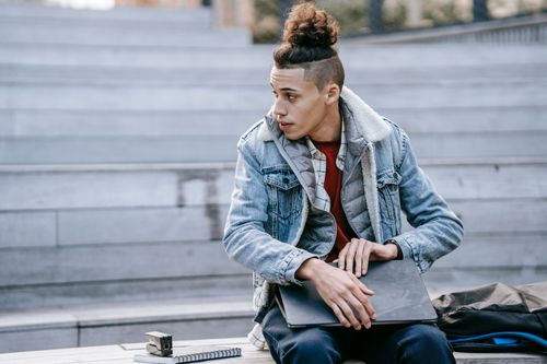 Free Young male with trendy hairdo looking into distance while sitting on wooden stairs with modern netbook during online work on blurred background Stock Photo