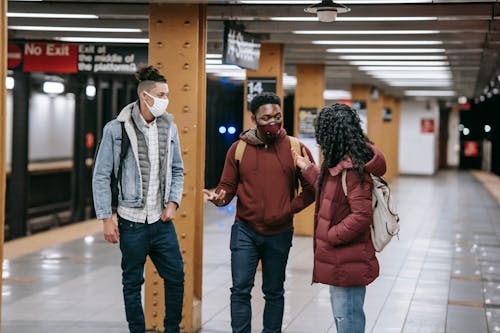 Multiracial group of friends with backpacks wearing protective masks looking at each other while standing on subway station during meeting