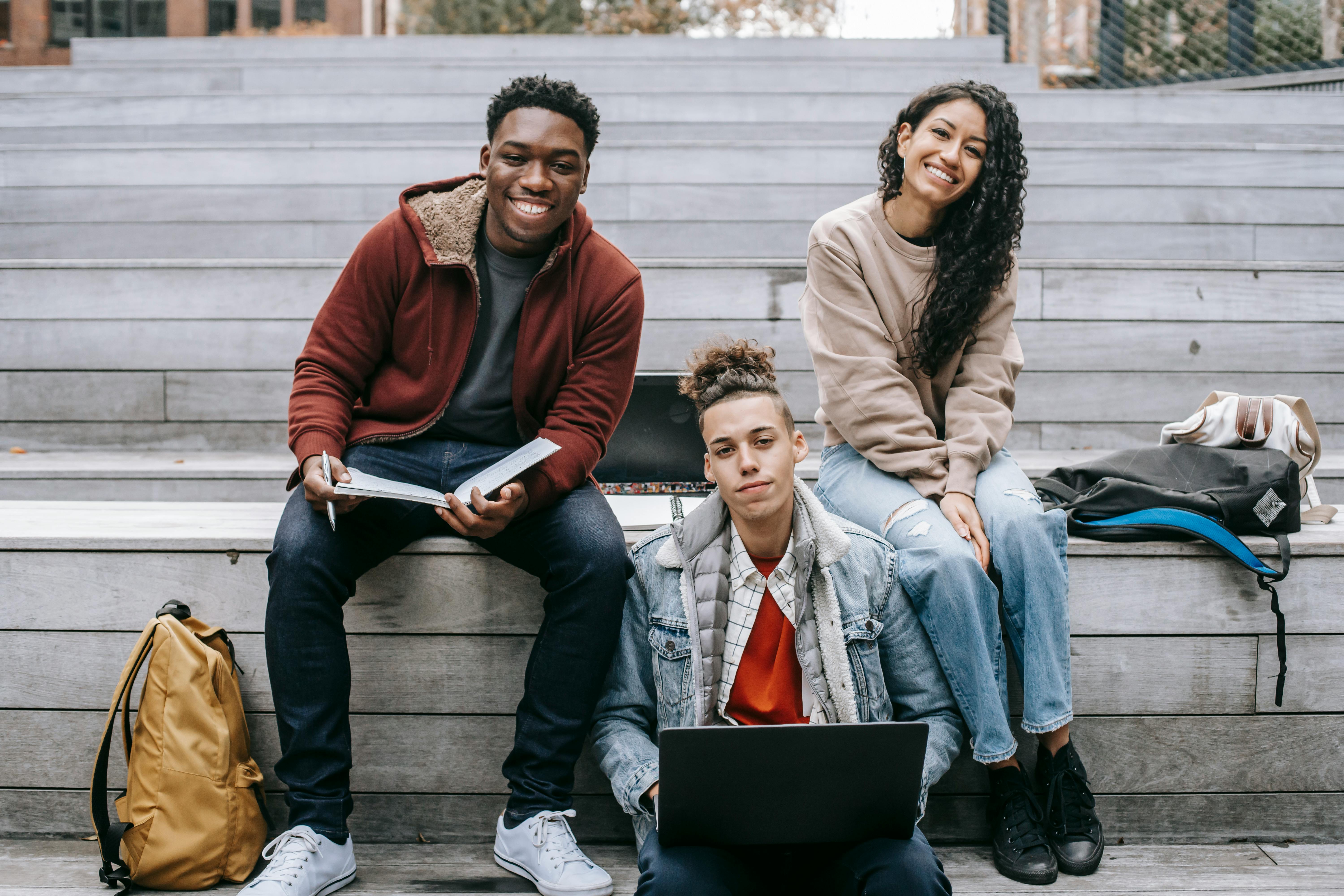 Three students sitting on outdoor steps with laptops and notebooks, smiling and studying together